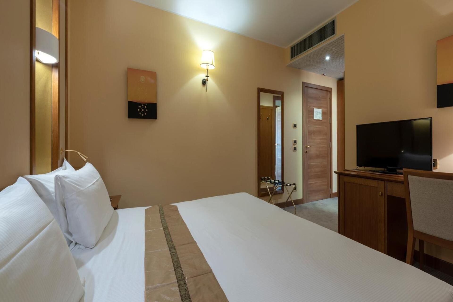 A hotel room with a bed, a desk with a television, a wall-mounted lamp, and a door leading to another room.