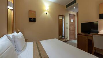 A hotel room with a bed, a desk with a television, a wall-mounted lamp, and a door leading to another room.