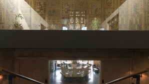Un hall d’hôtel doté d’un plafond haut, de motifs décoratifs élaborés sur les murs et d’un escalier en verre menant à un niveau inférieur.