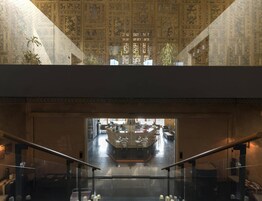 Un vestíbulo de hotel con techo alto, detalles ornamentales en las paredes y una escalera de cristal que desciende a un nivel inferior.