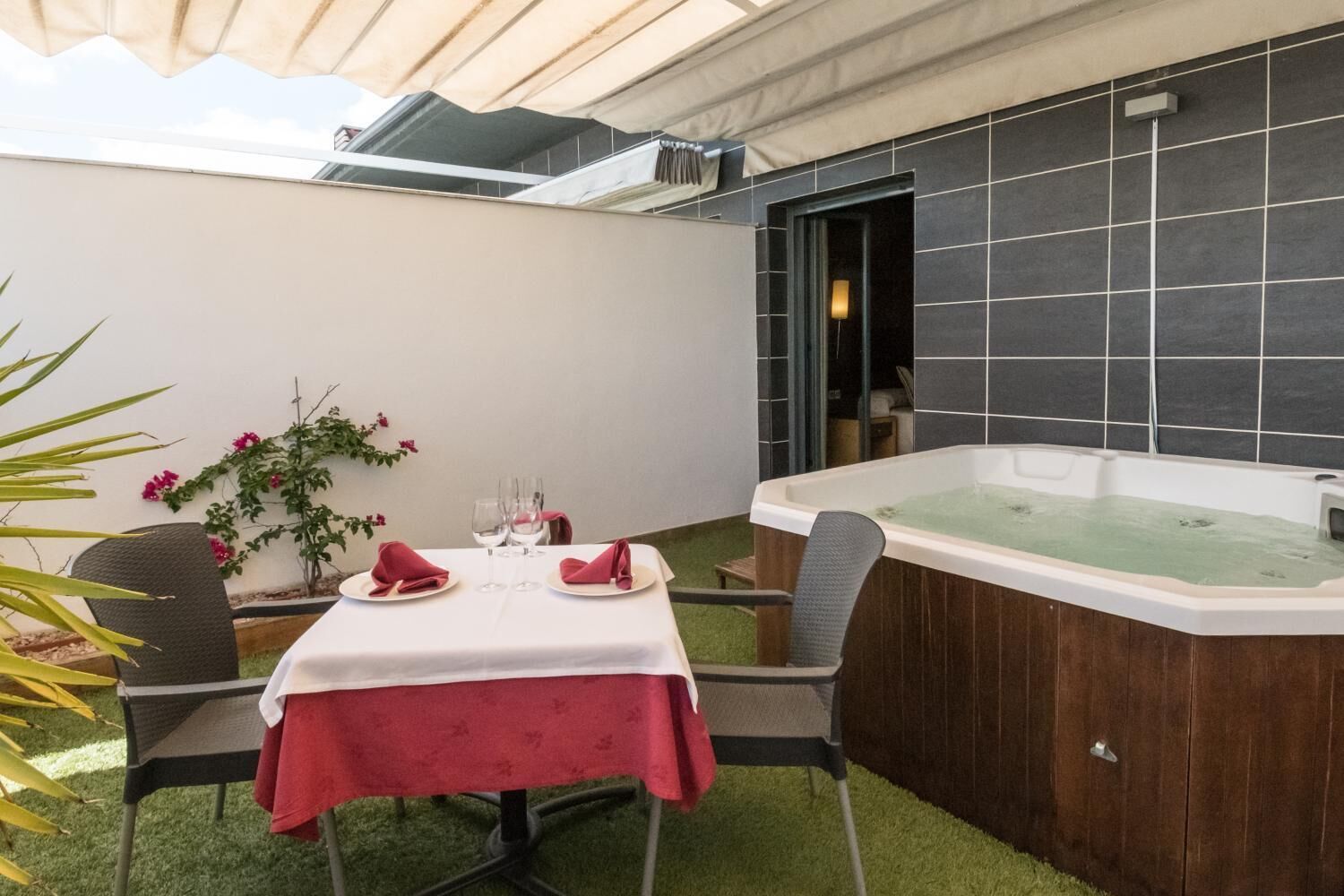 A patio area with a table set for two, a hot tub, and a small garden with pink flowers.