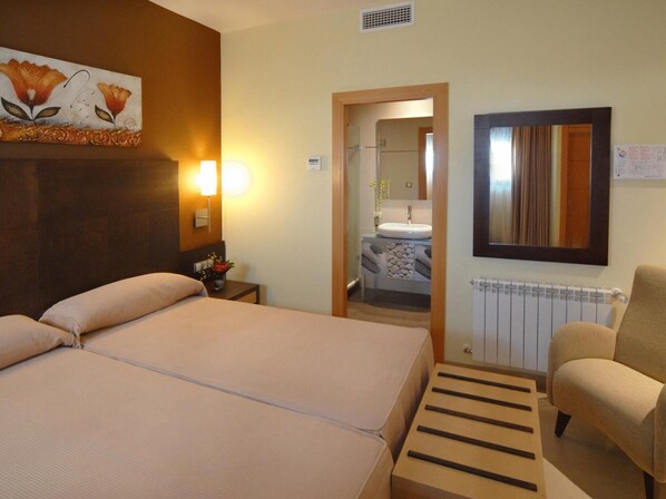 Habitación de hotel con cama doble, lámparas de noche, una mesita, una silla, un baño visible a través de una puerta abierta y un radiador.