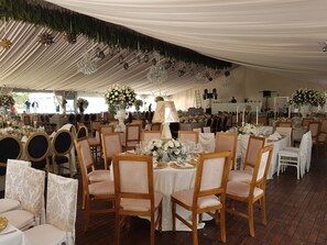 Un espace événementiel aménagé sous une tente, avec des tables rondes, des chaises en bois et des centres de table floraux.