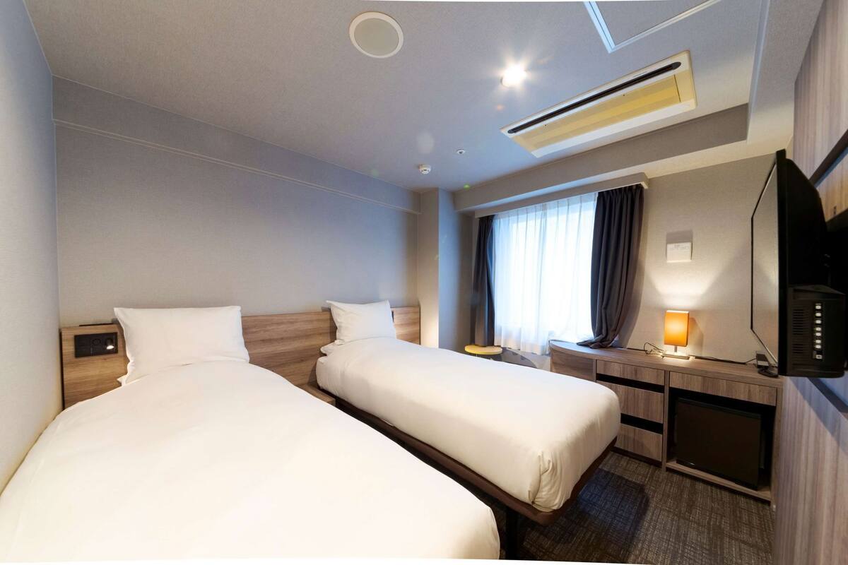 A hotel room with two beds, a desk, a TV, and a window with curtains.
