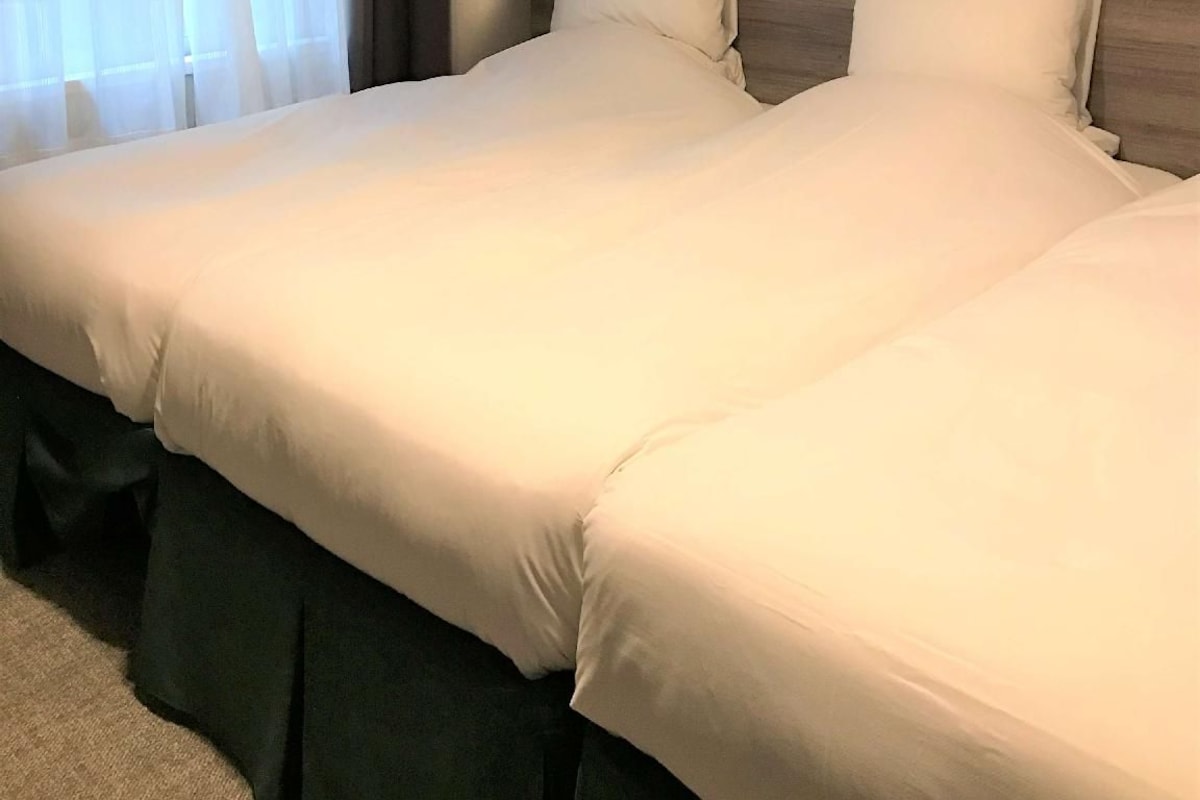 Habitación de hotel con dos camas, una ventana con cortinas, una mesita de noche y un aplique de pared.