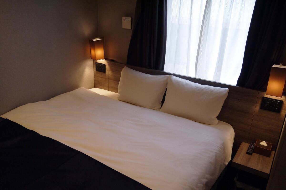 A neatly made bed with white linens, two bedside lamps, a wooden headboard, and a window with curtains.
