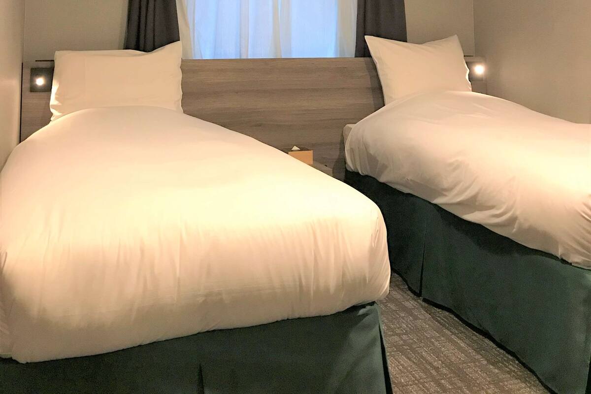 A hotel room with two single beds, a window with curtains, an air conditioning unit, and a night light.