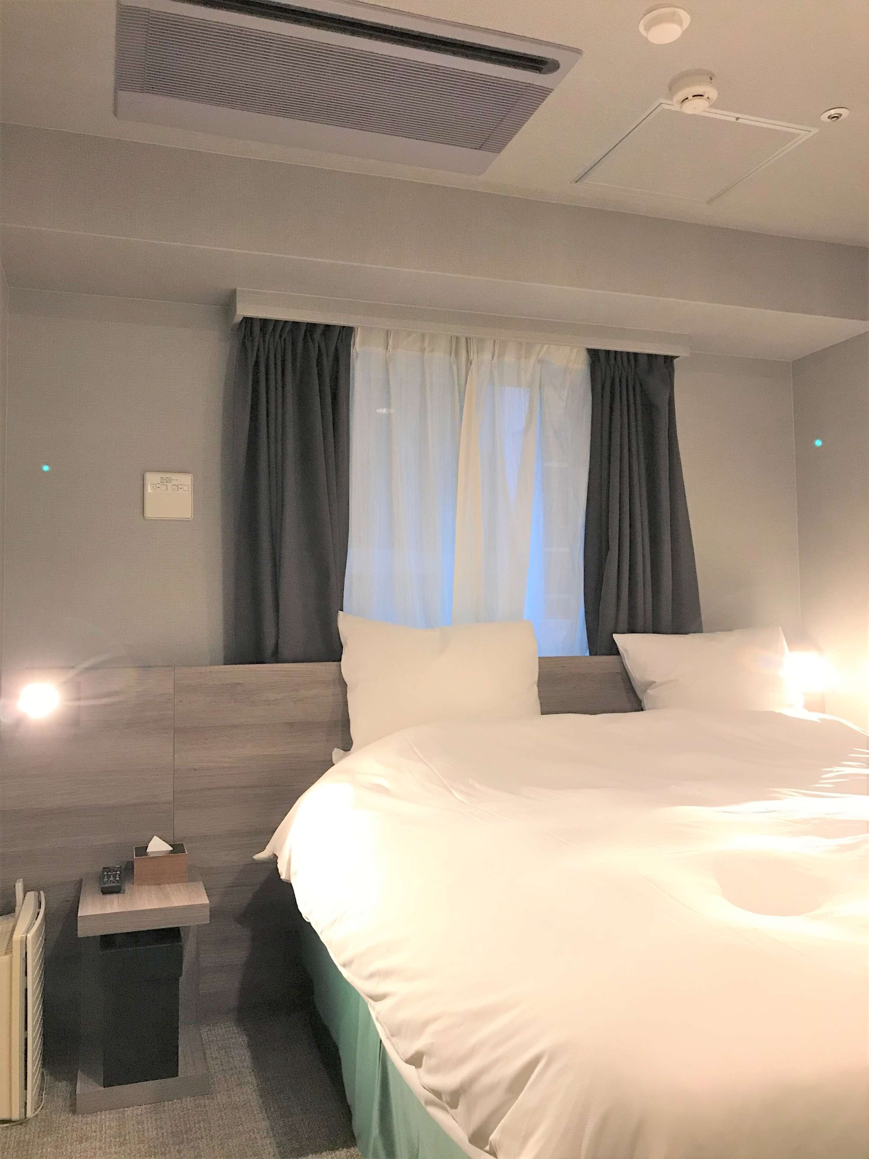 Ein Hotelzimmer mit einem großen Bett, einem Nachttisch mit Lampe, einem Fenster mit Vorhängen und einem Deckenventil.