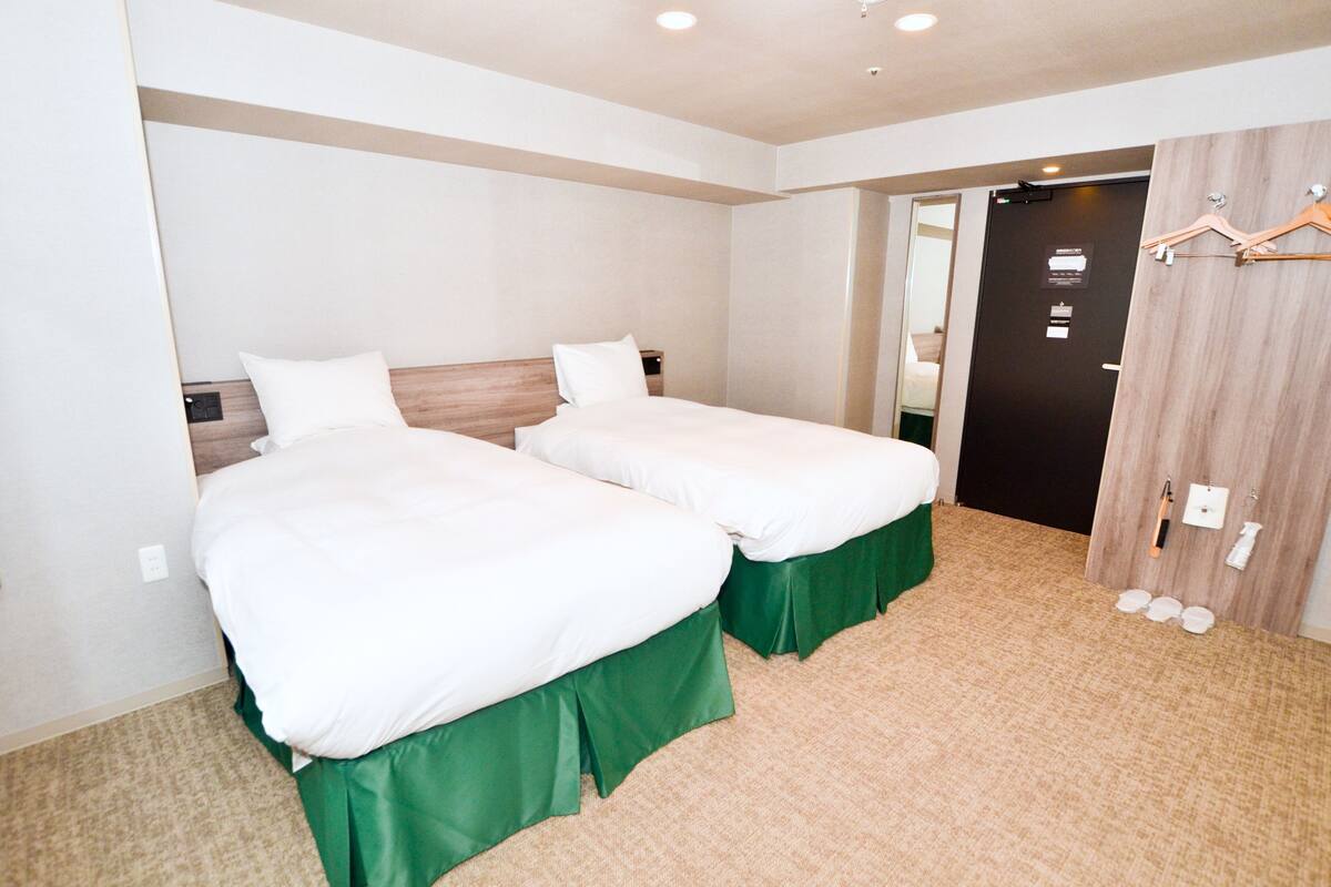 A hotel room with two beds, green bed skirts, a wooden headboard, a wooden wardrobe, and a door with a mirror.
