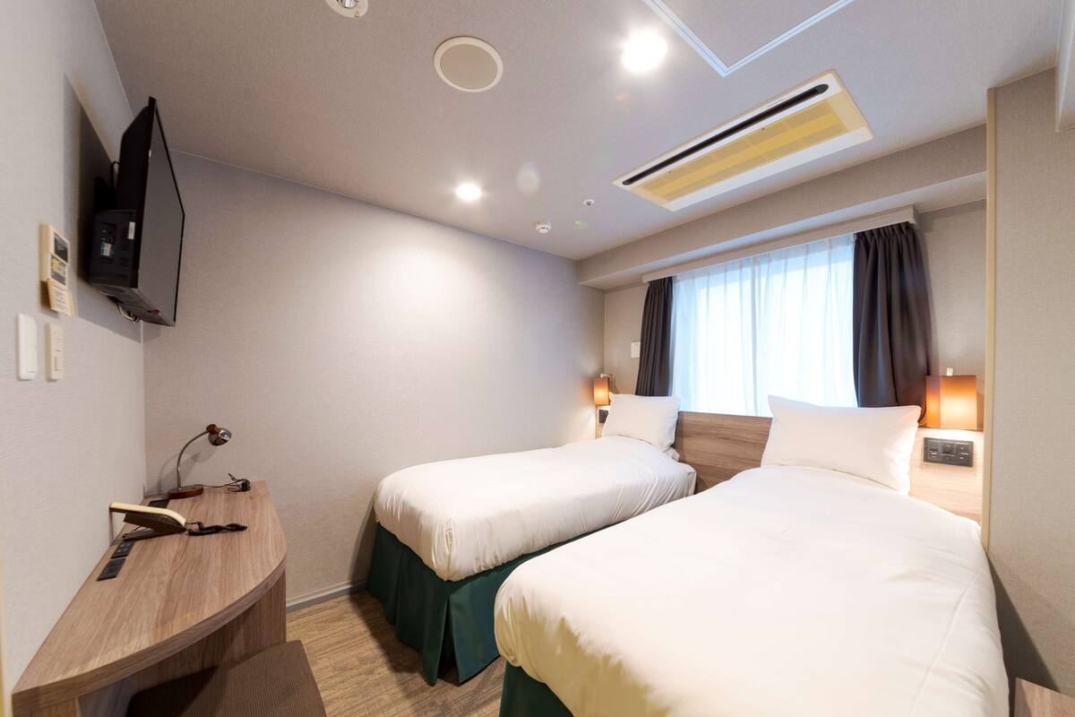 A hotel room with two single beds, a desk, a TV, and a window with curtains.
