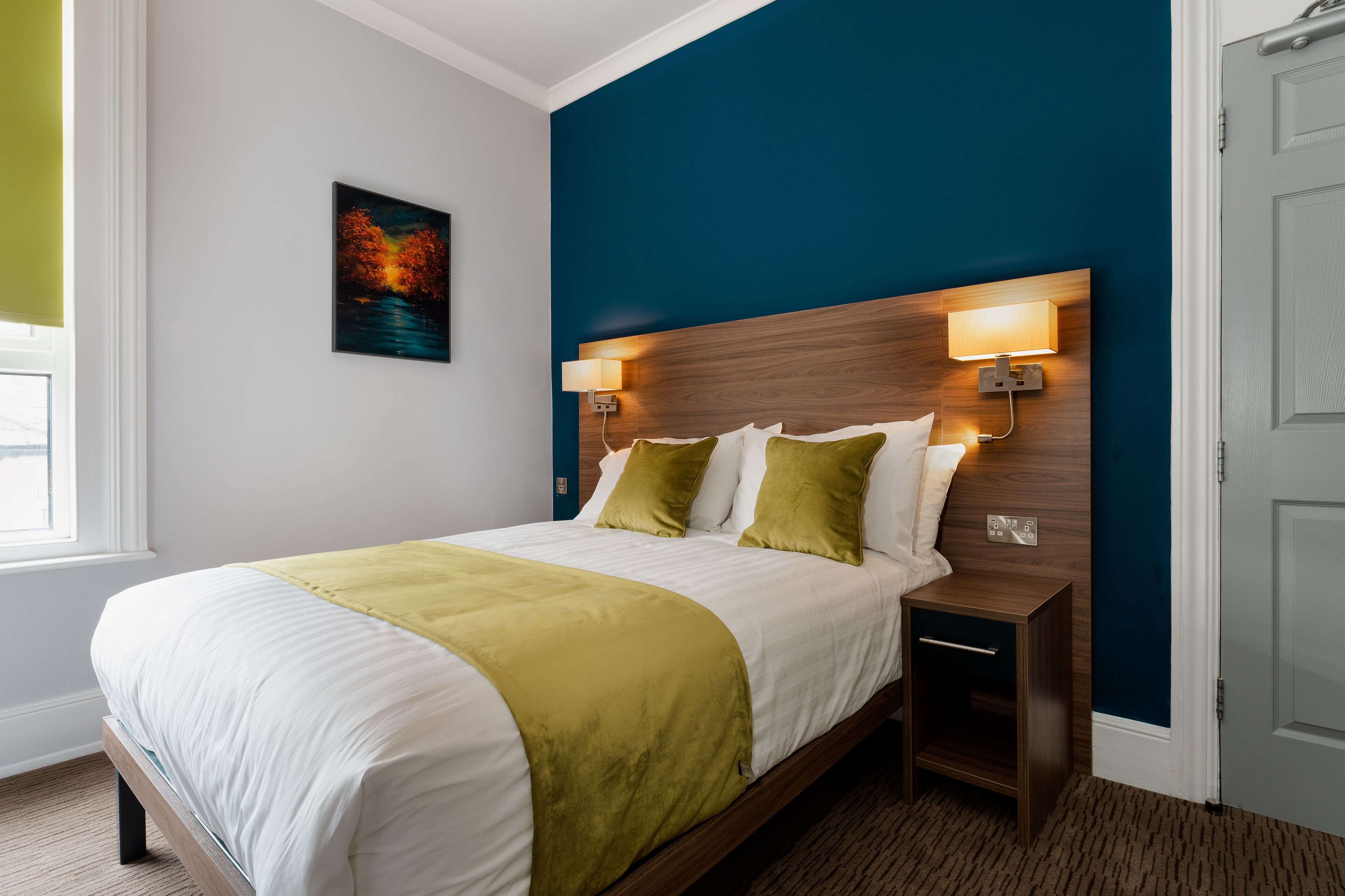 Cama bem arrumada com manta verde, duas luminárias de cabeceira e mesa de apoio.