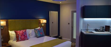 A modern hotel room with a large bed, a bedside table, a nightstand, a built-in desk with a sink, and a mirror above the desk.