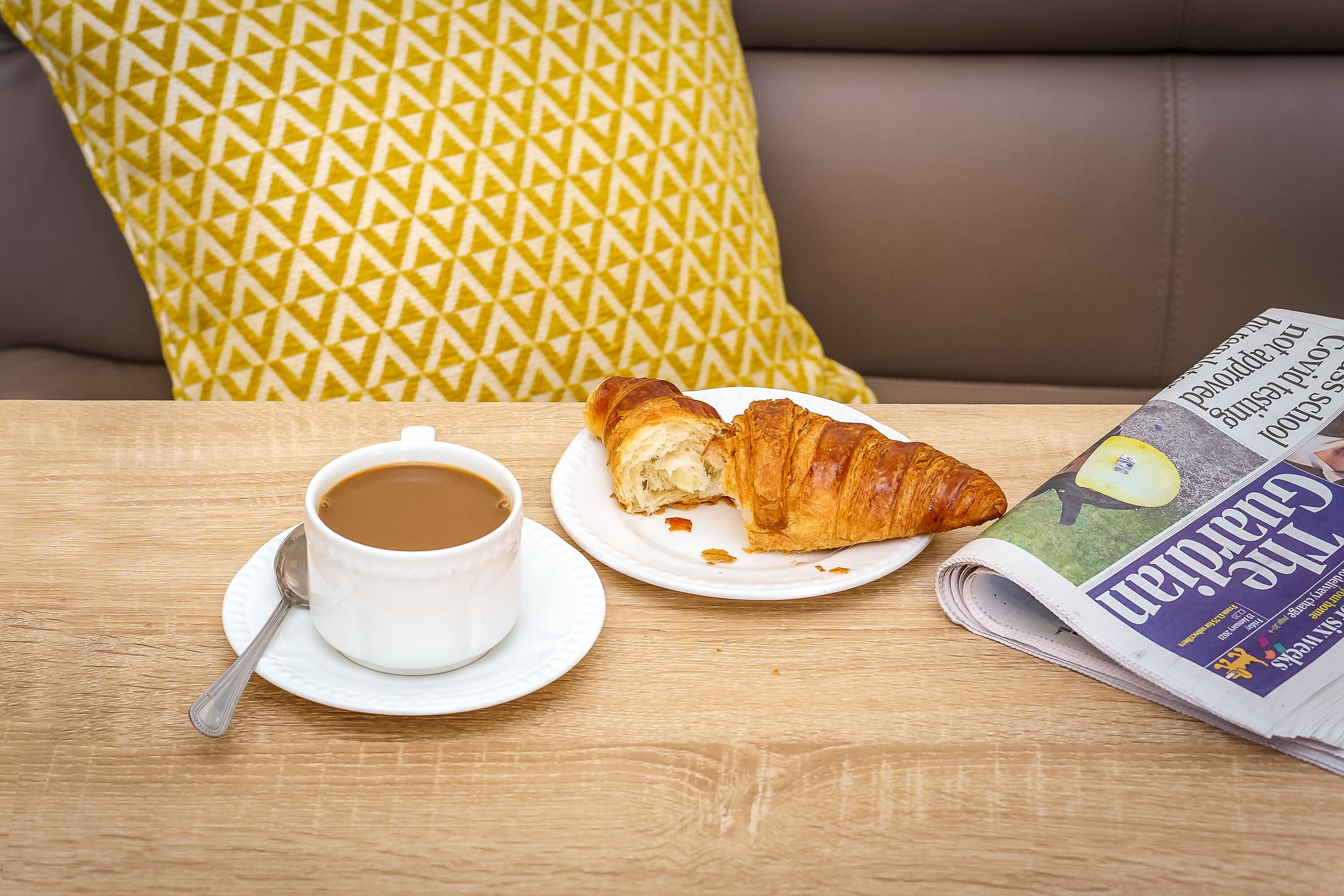 Un desayuno con una taza de café, un croissant y un periódico sobre una mesa de madera.