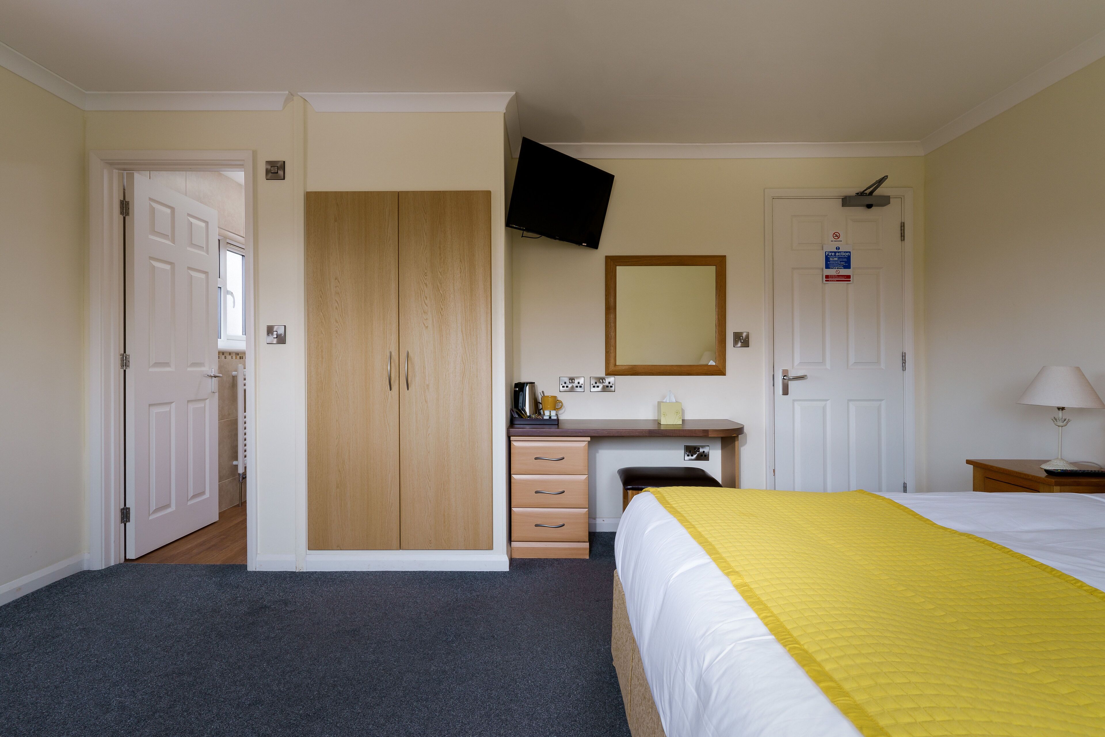 Una habitación de hotel con cama, escritorio con silla, armario, televisor montado en la pared y una puerta.