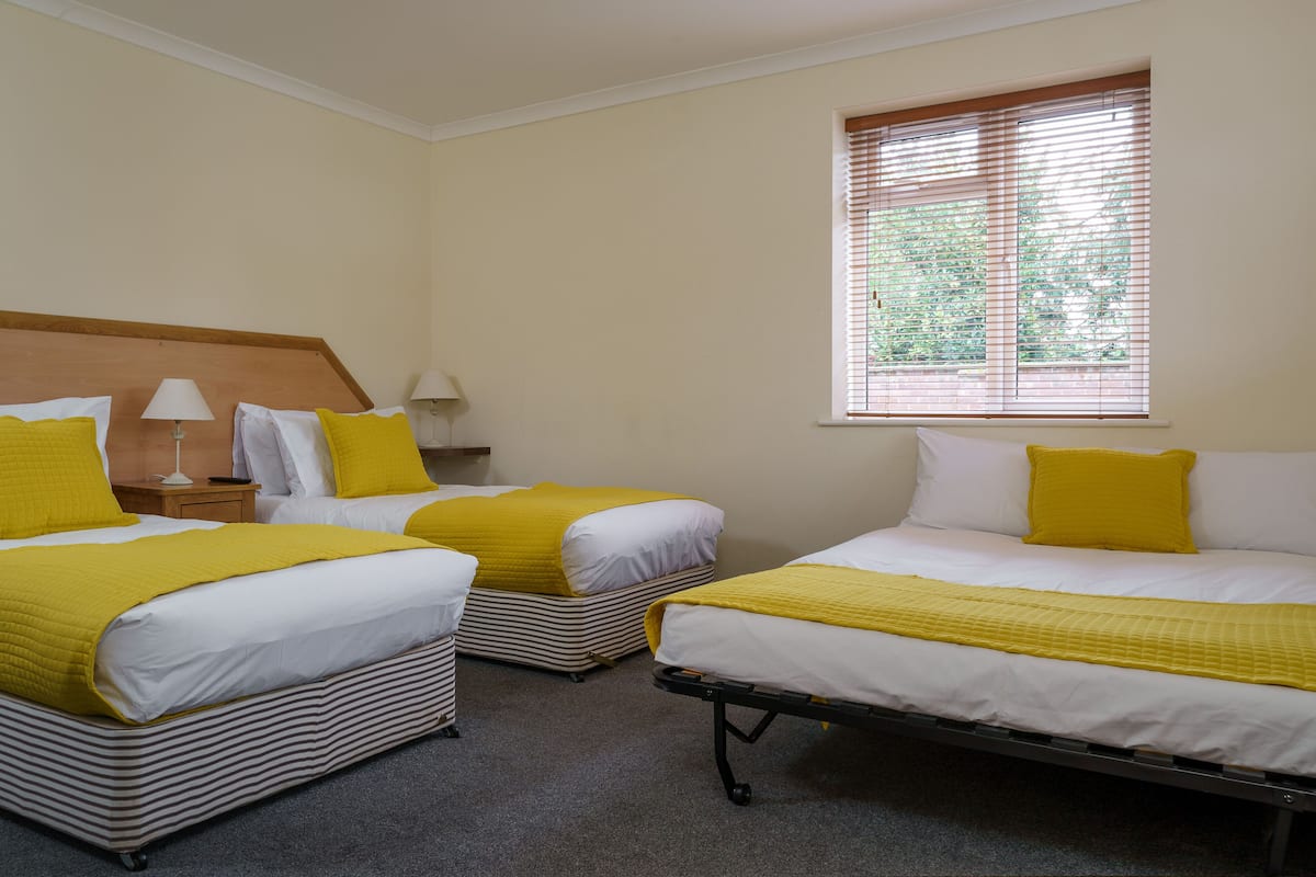 Habitación de hotel con dos camas, cada una con una manta amarilla y almohadas blancas, cabecera de madera y una ventana con persianas.