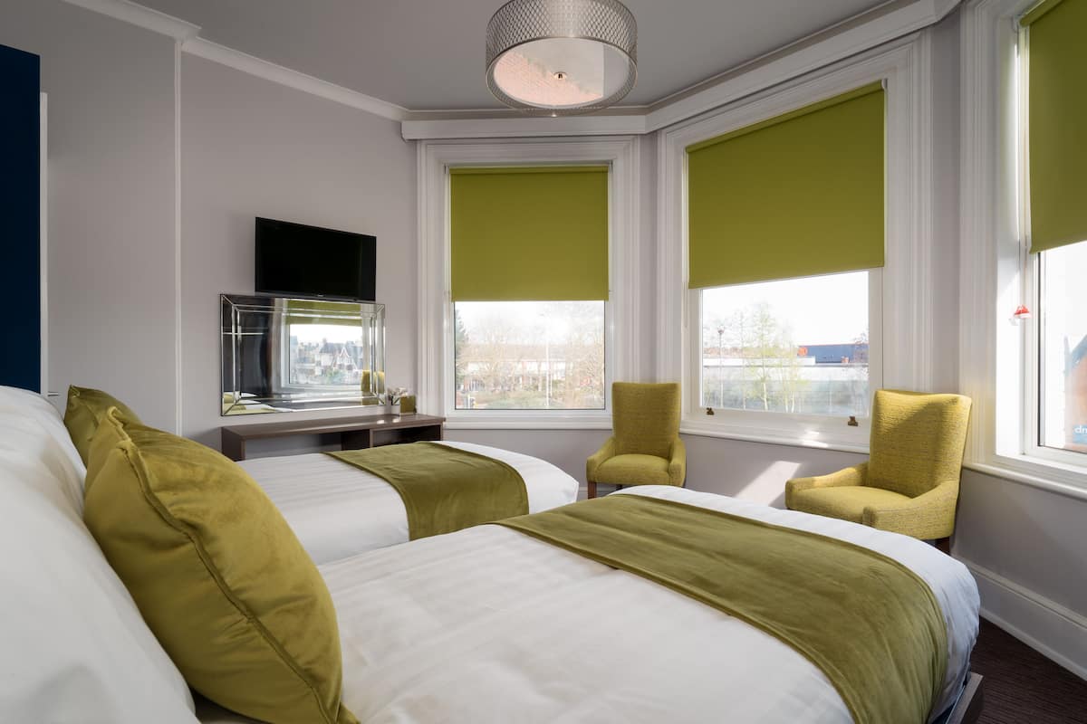 Habitación de hotel con dos camas, una silla, un televisor y ventanas amplias con persianas.