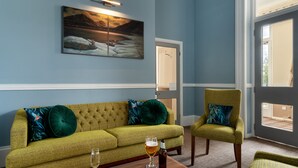 Una habitación con un sofá verde, un sillón verde, una cerveza en un vaso y un cuadro en la pared.