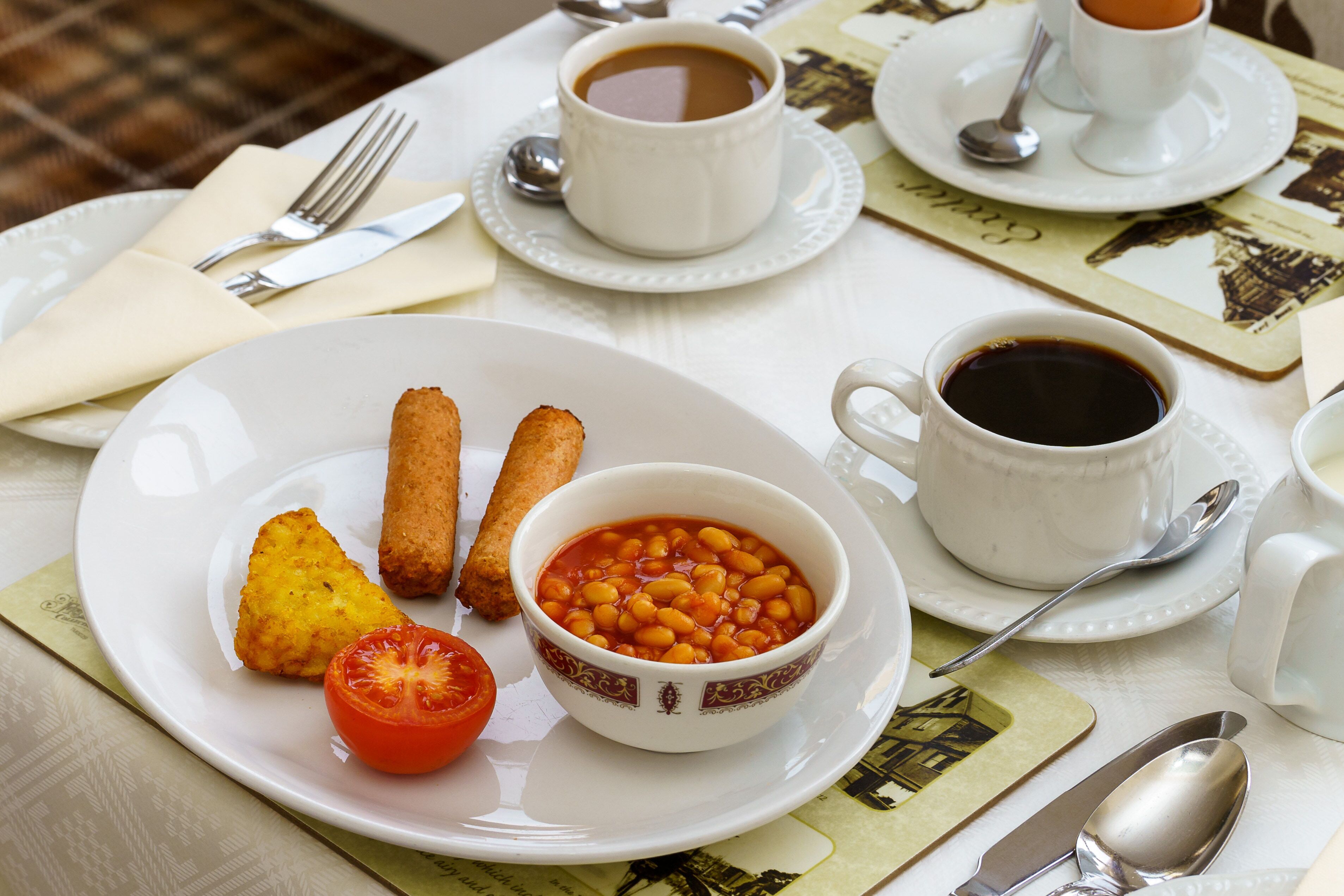 Ein Tisch ist gedeckt mit einem Teller Essen, einer Bohnenschale, einer Tomate und zwei Kaffeetassen.