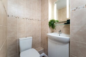 Un baño con baldosas color beige, un inodoro blanco, un lavabo con espejo y una planta pequeña sobre el lavabo.