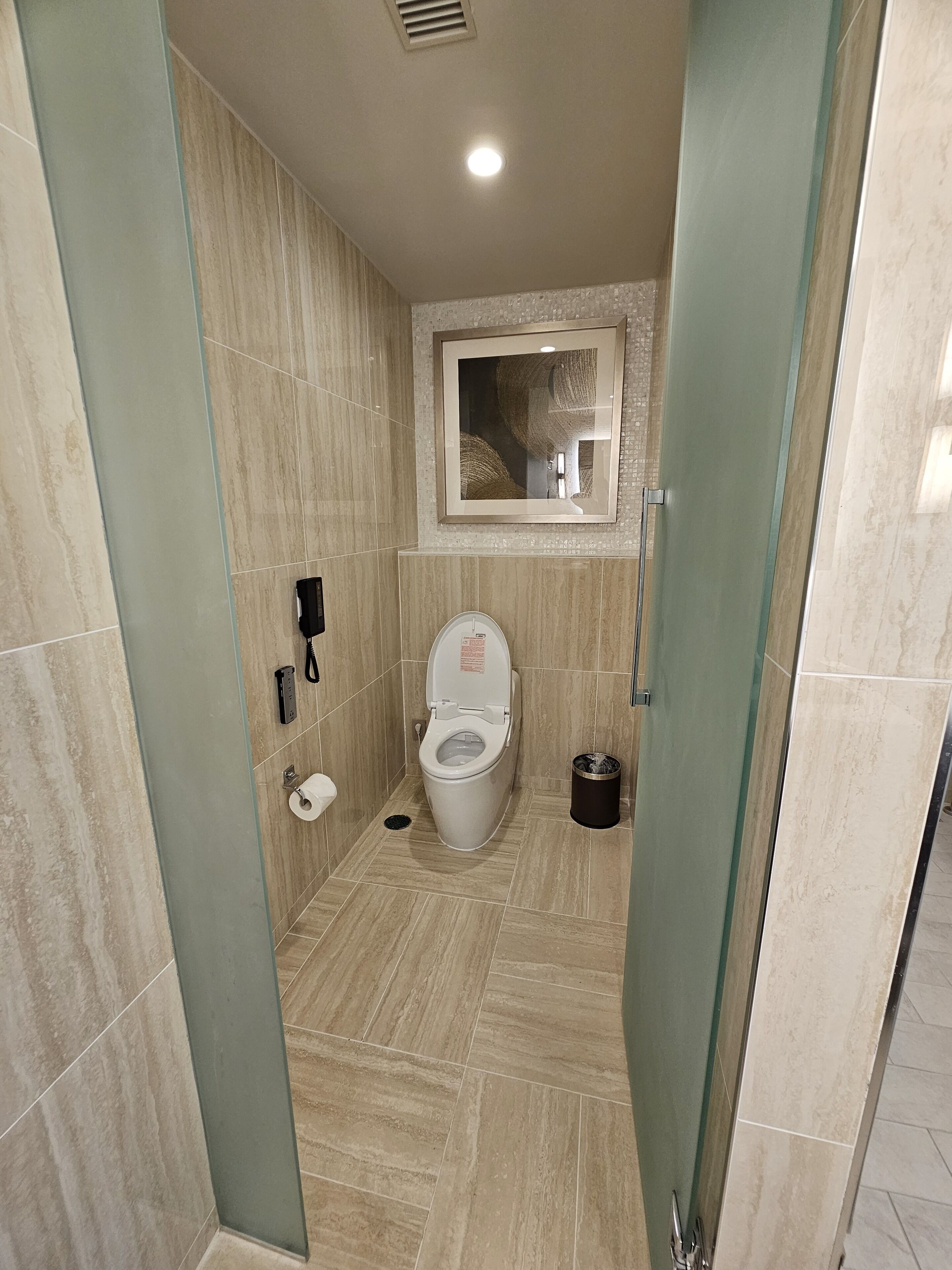 A modern bathroom with a toilet, a trash bin, and a wall-mounted phone.
