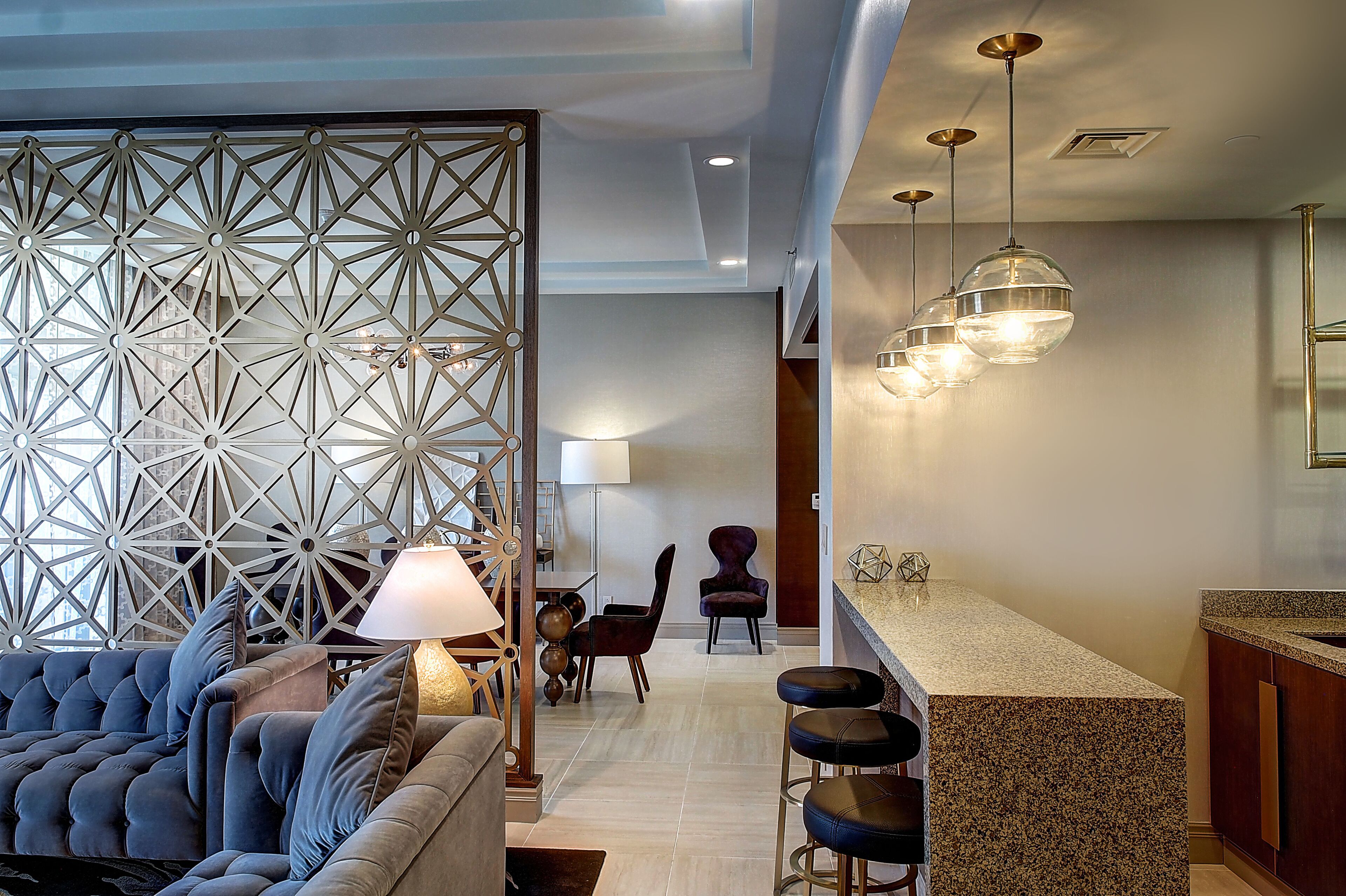 A modern interior with a geometric wall design, a seating area with a sofa and chairs, and a kitchen with bar stools and pendant lighting.