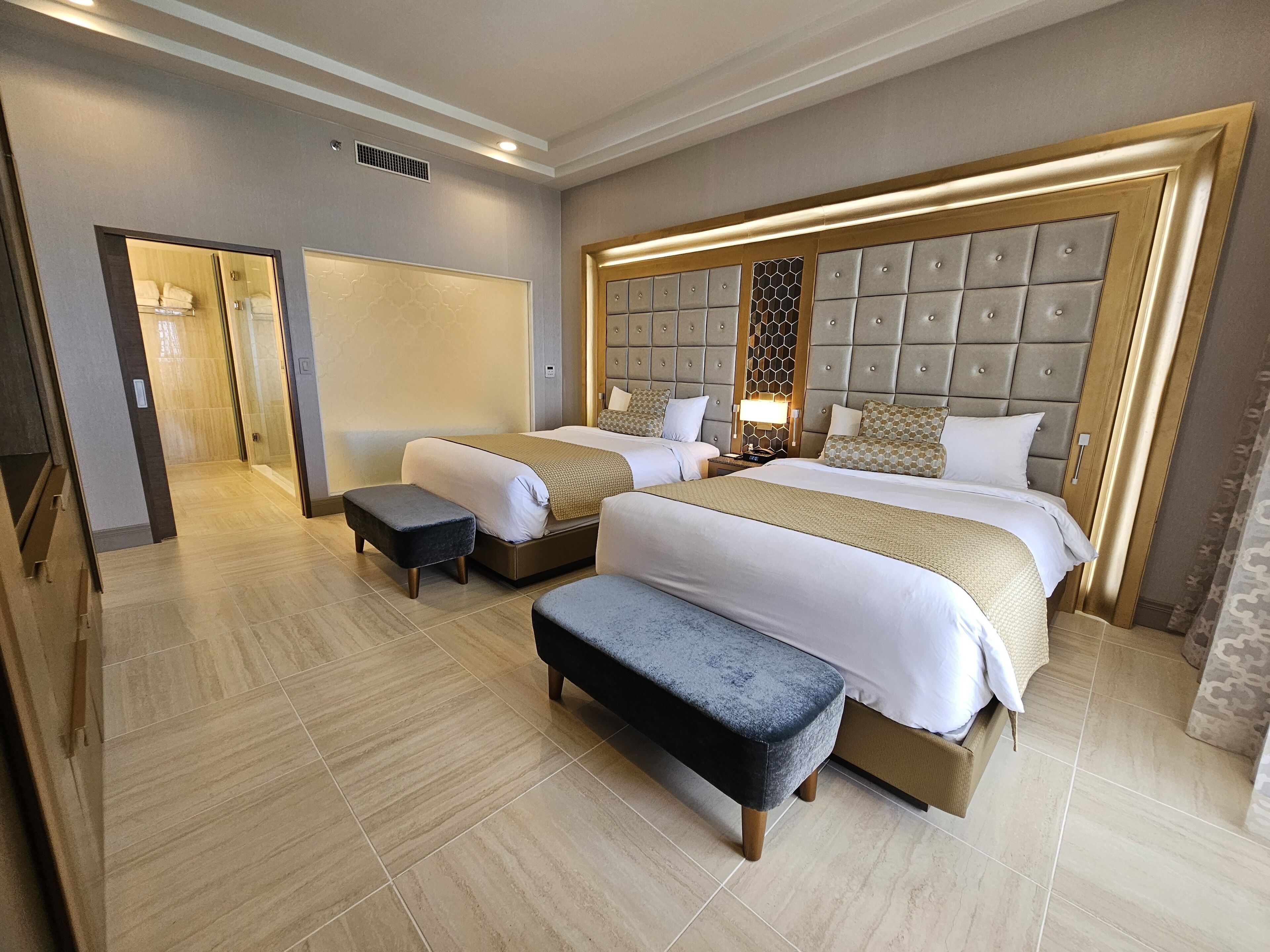 Quarto de hotel com duas camas, cabeceira grande acolchoada, um banco e um closet espaçoso.