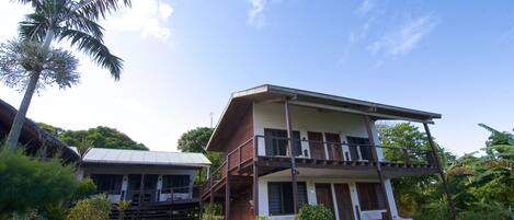 A two-story house with a balcony, surrounded by lush greenery and a palm tree.