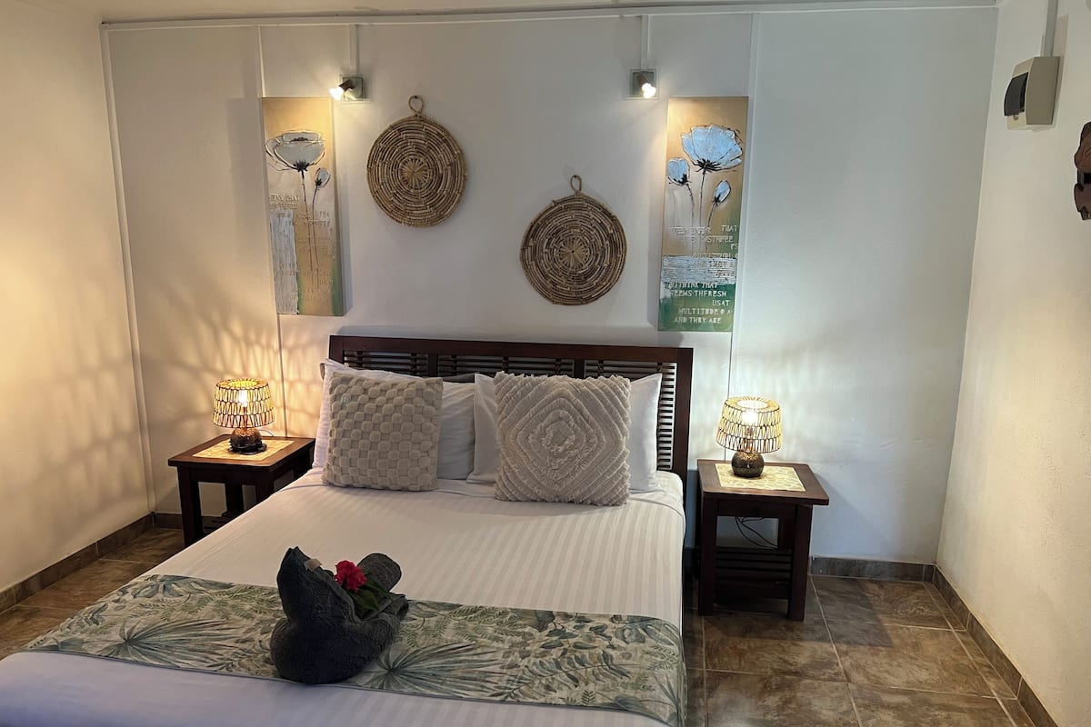 Una cama cuidadosamente tendida con una colcha de motivos florales y almohadones decorativos, dos lámparas de noche y arte en la pared que incluye piezas tejidas circulares y una cita enmarcada.