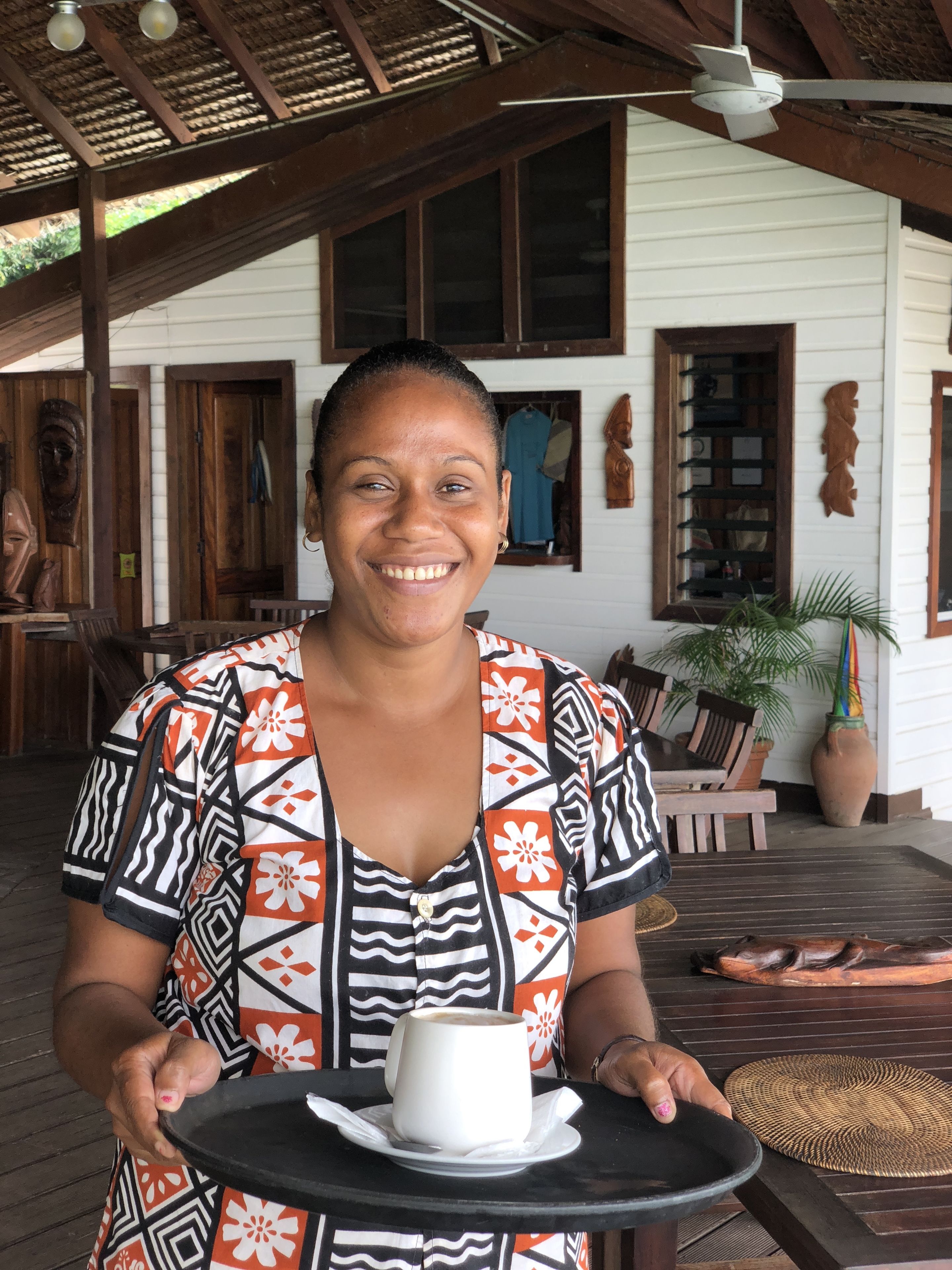 A person holding a cup of coffee on a table in a covered outdoor area with wooden furniture and a white building in the background.