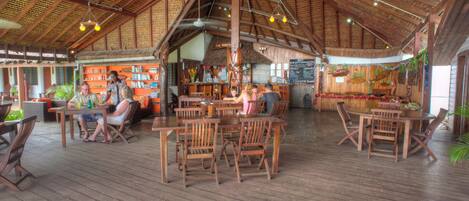 An indoor wooden deck with tables and chairs, a bar area, and a thatched roof.