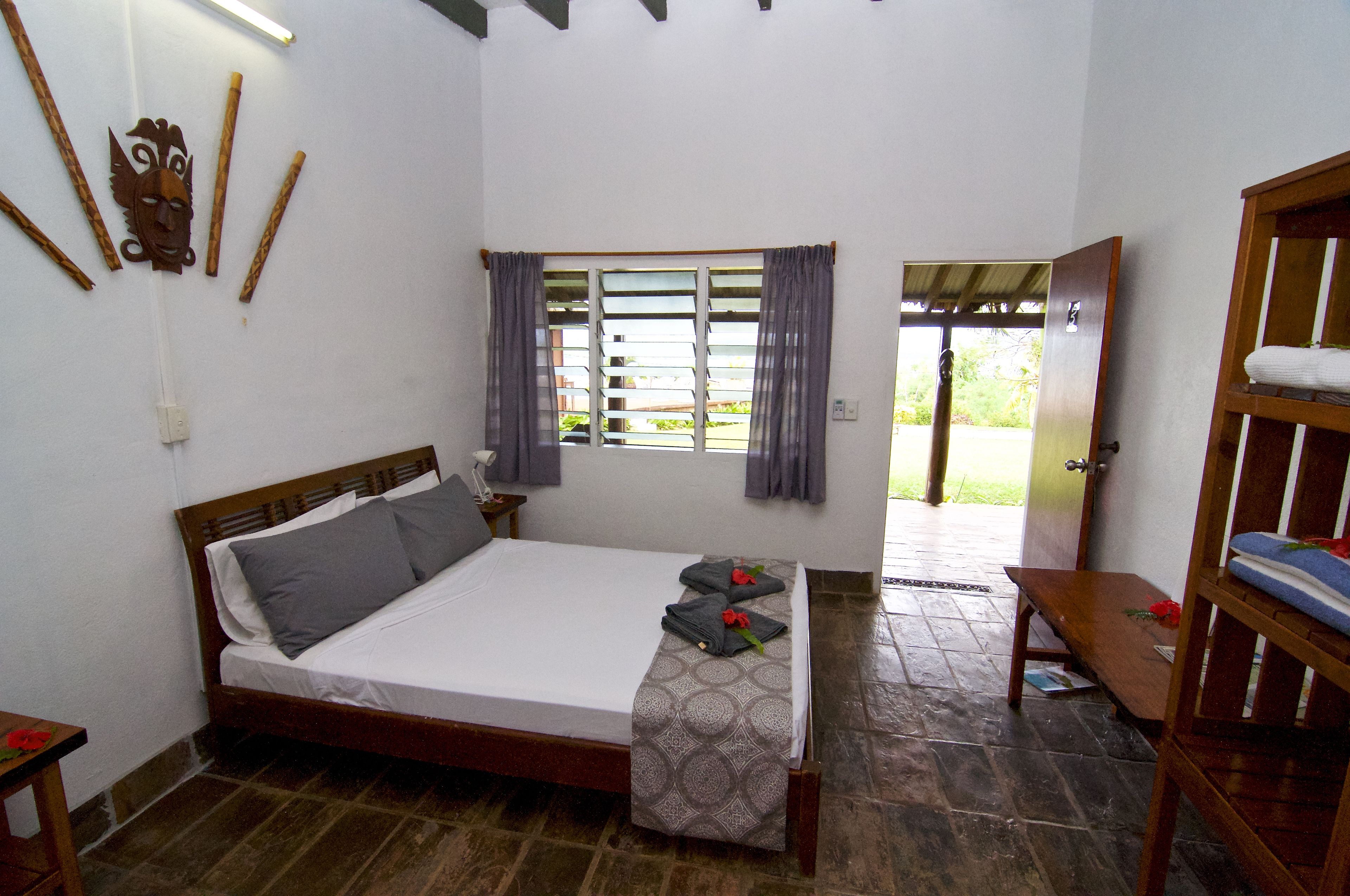 A bedroom with a bed, a bench, a wooden wardrobe, a window with curtains, and a door leading outside.
