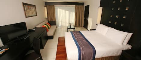 Habitación de hotel con una cama grande, un escritorio con televisor, una silla y un sofá con cojines de colores.