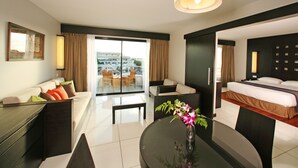 A modern hotel room with a bed, a sofa, a dining table, and a balcony with a view of buildings.