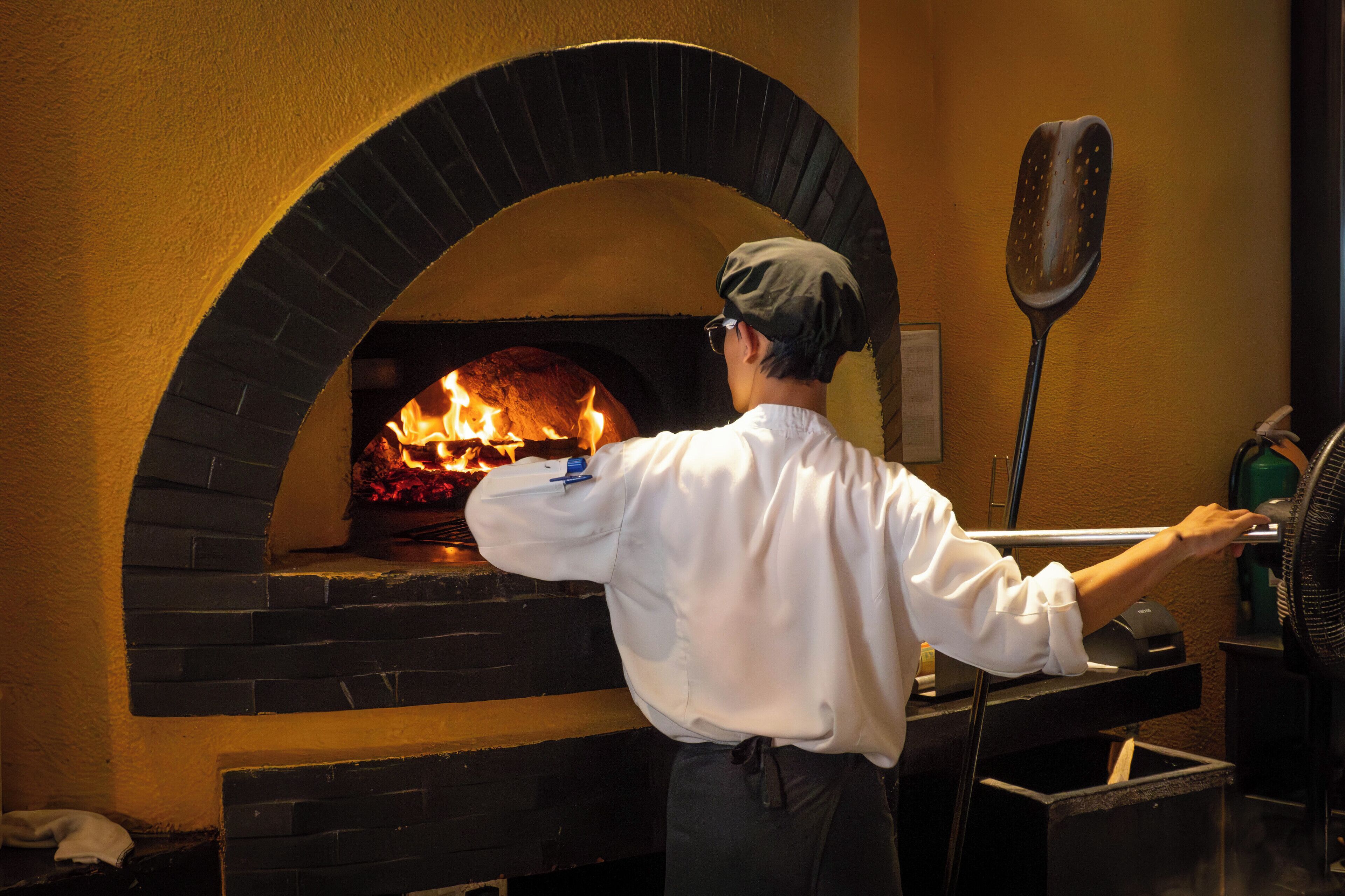 Un cocinero con camisa y delantal blanco frente a un horno de ladrillo con llamas, que indica un horno para pizza.