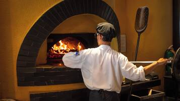 Un cocinero con camisa y delantal blanco frente a un horno de ladrillo con llamas, que indica un horno para pizza.