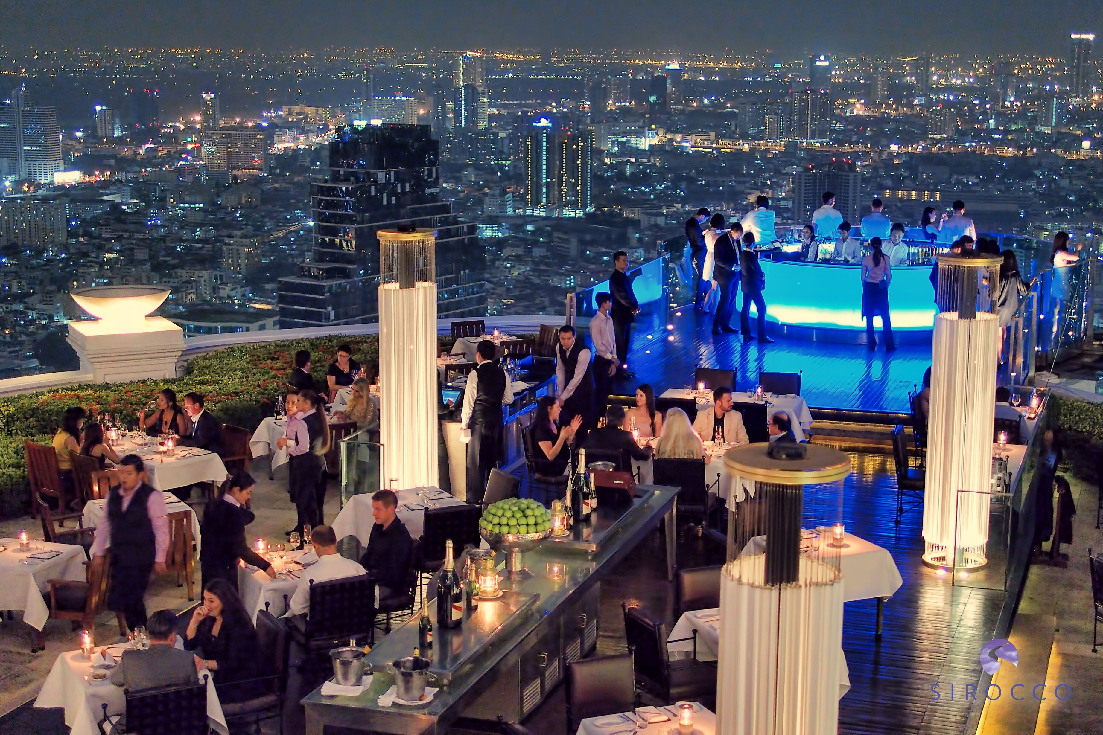 A rooftop bar with tables and chairs, a stage for performances, and a cityscape view in the background.