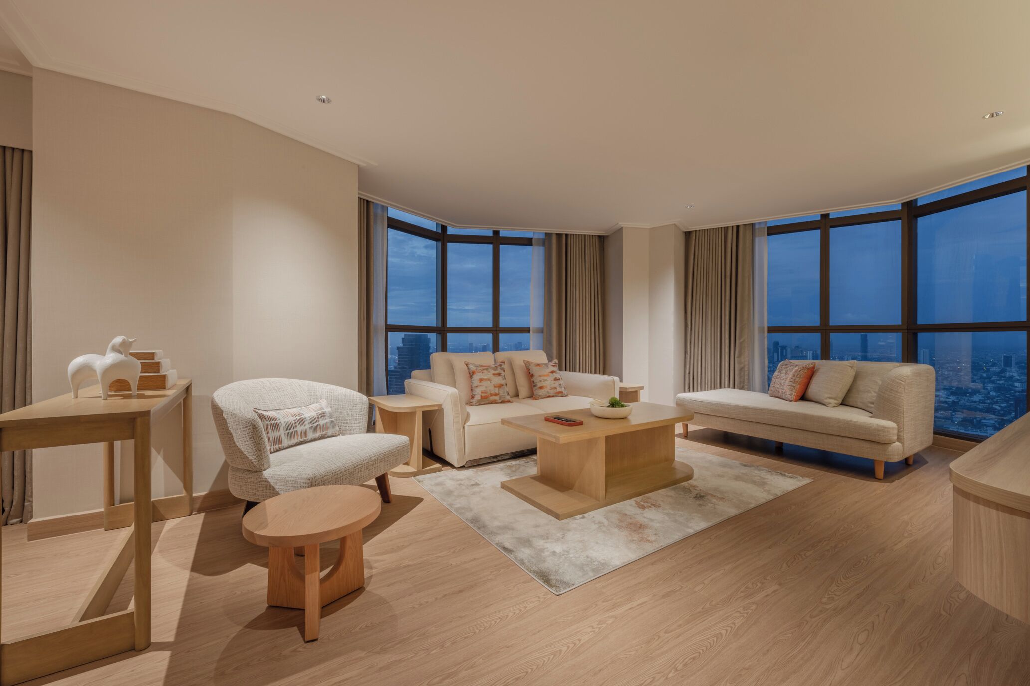 A modern living room with a sofa, coffee table, and side tables, large windows with curtains, and a city view.