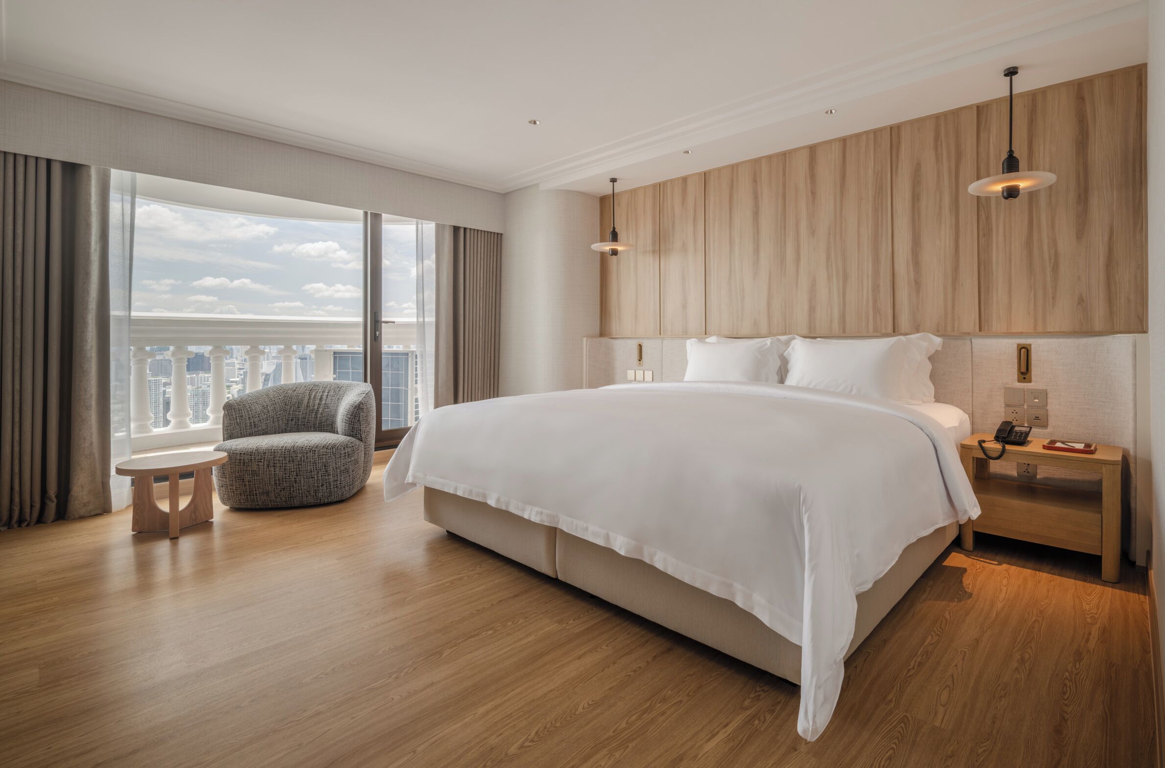A modern hotel room with a large bed, a round chair, a nightstand with a phone, and a view of the city.