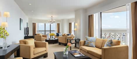 Un salón espacioso con sofás color beige, una mesa de centro y un televisor. Amplias ventanas ofrecen vistas a la ciudad y al mar.
