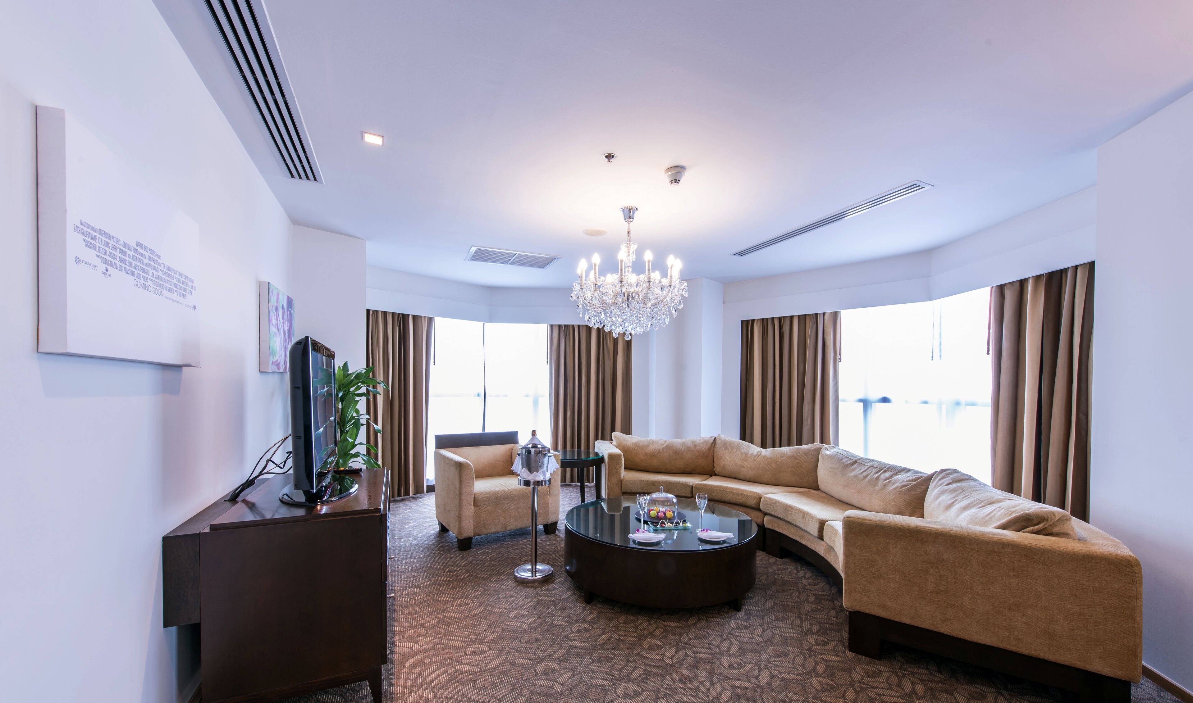 A modern hotel room with a large beige sofa, a glass coffee table, a TV, and a chandelier.
