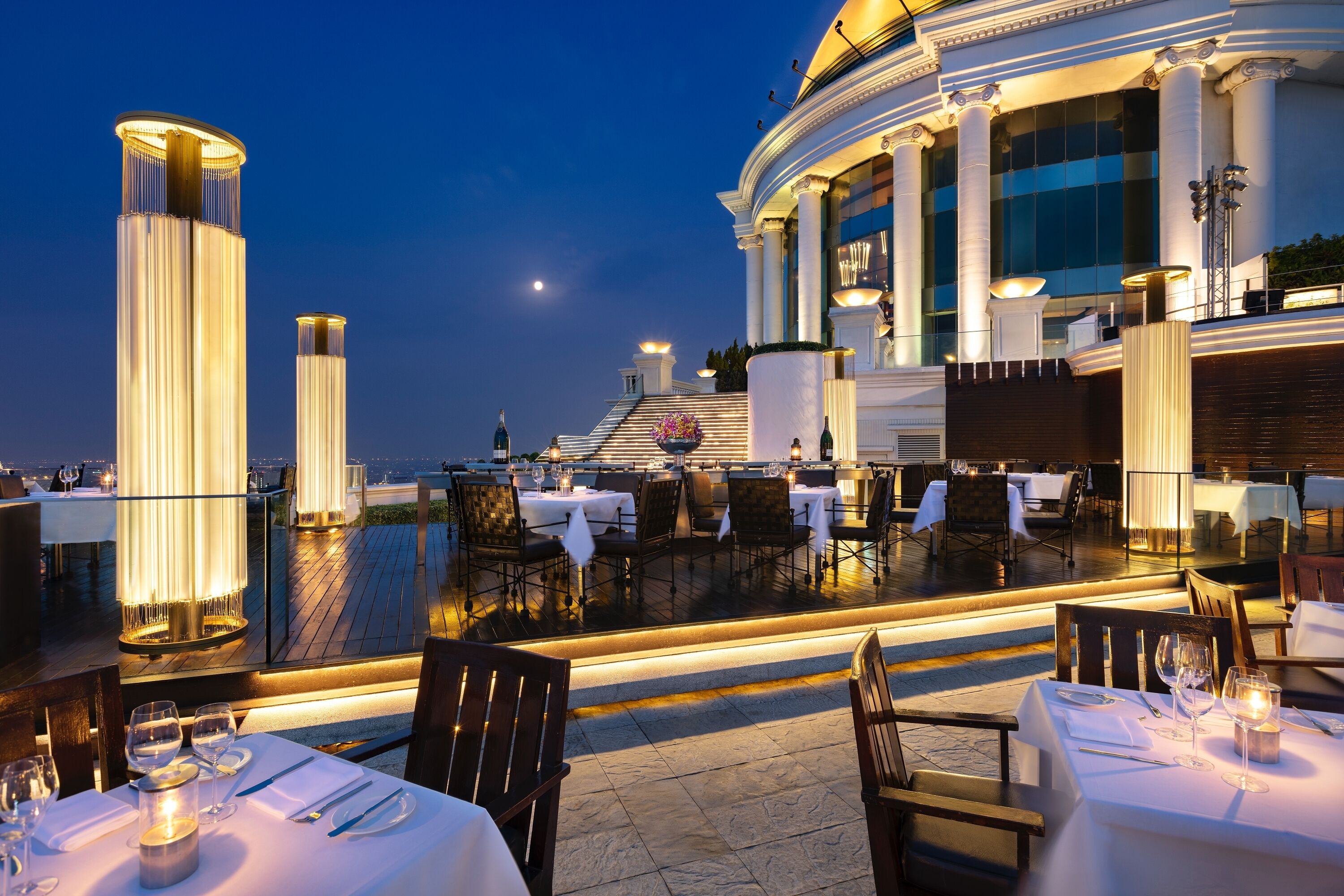 An outdoor dining area with tables set for dinner, a large illuminated column, and a view of the city skyline at dusk.
