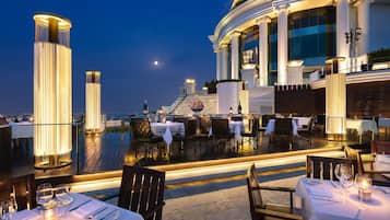 An outdoor dining area with tables set for dinner, a large illuminated column, and a view of the city skyline at dusk.