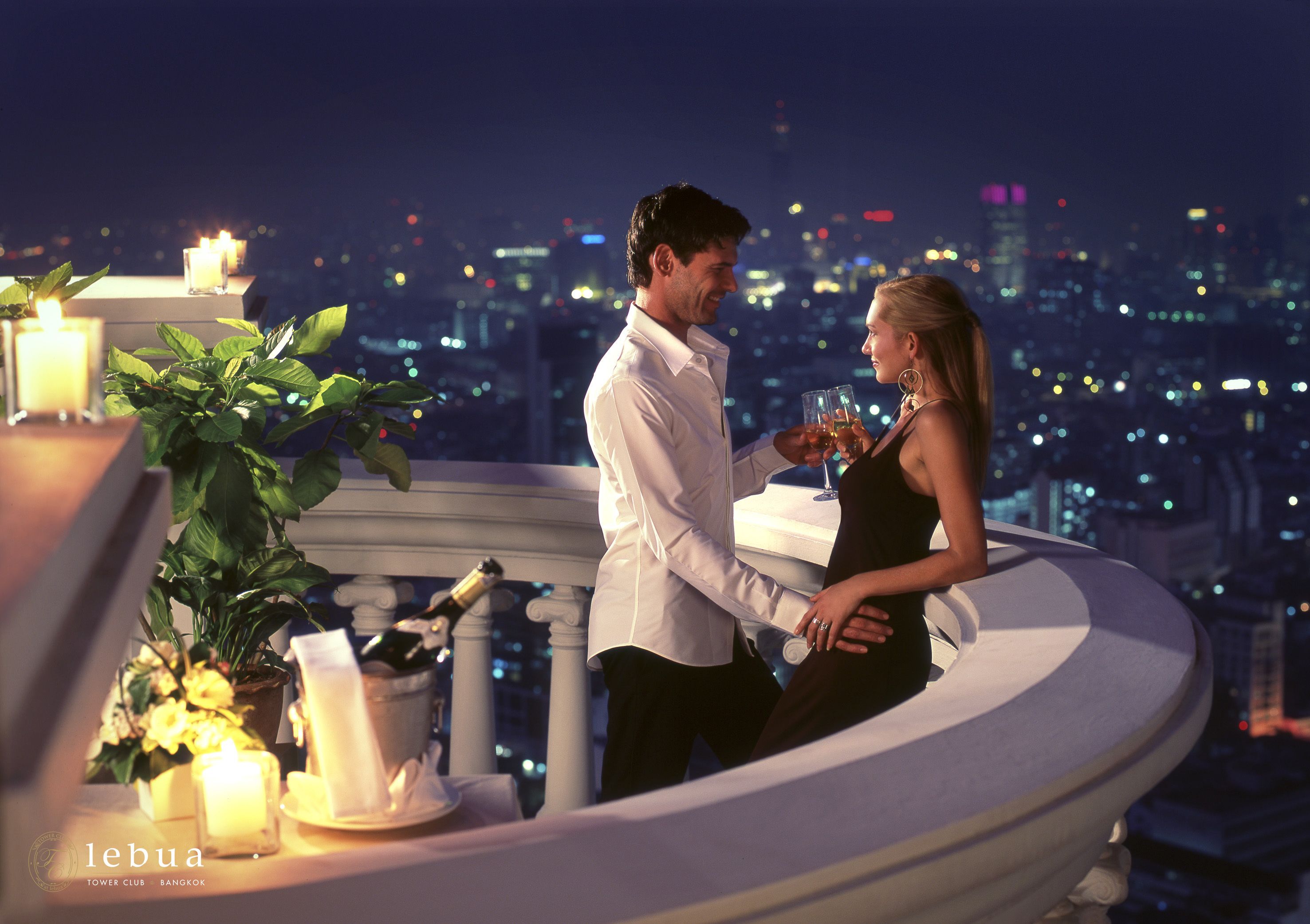 A couple on a balcony with a cityscape view, candles, and a potted plant.