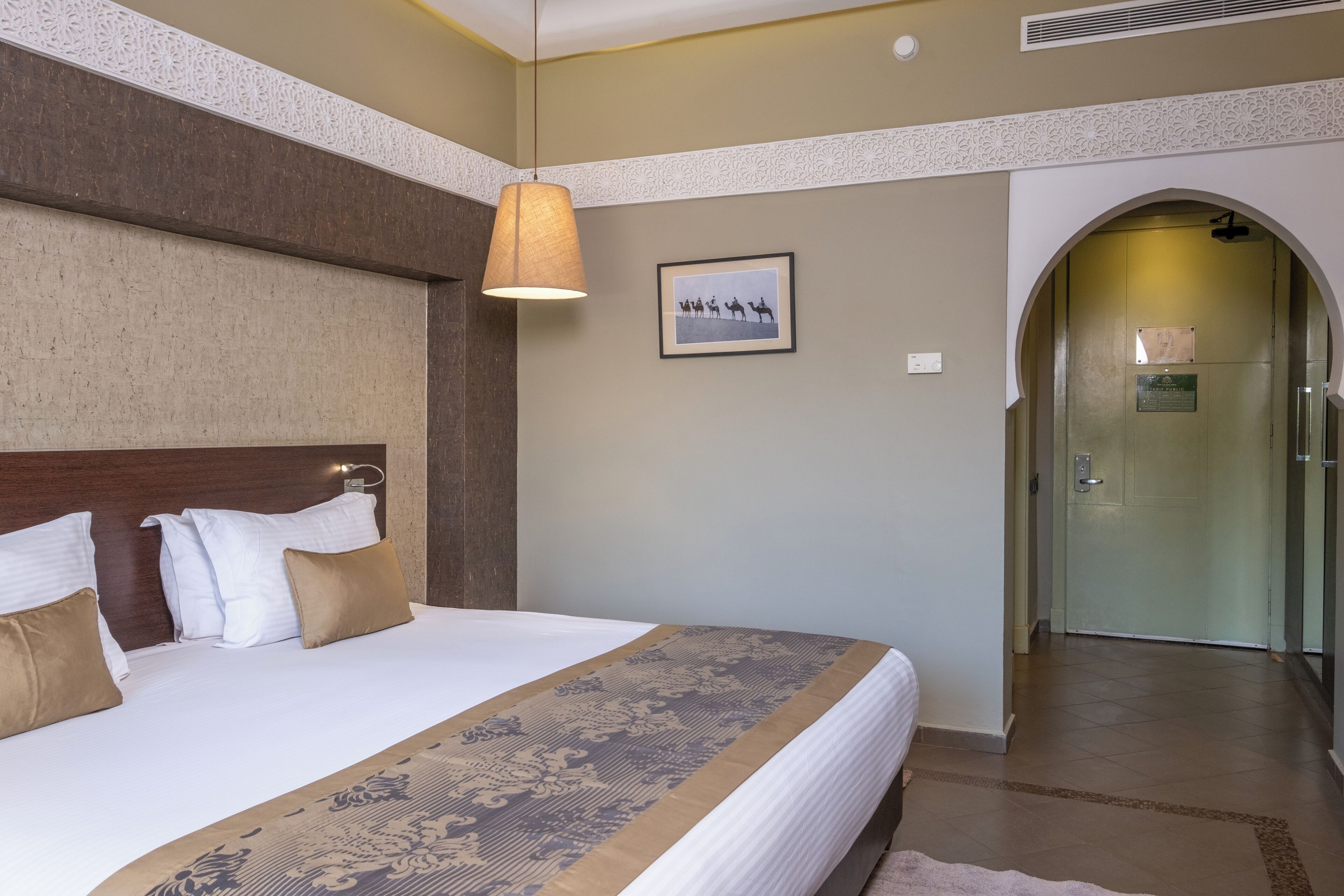 Habitación de hotel con una cama grande, un cabecero, una lámpara colgante, un cuadro en la pared y un arco que da a otra habitación.