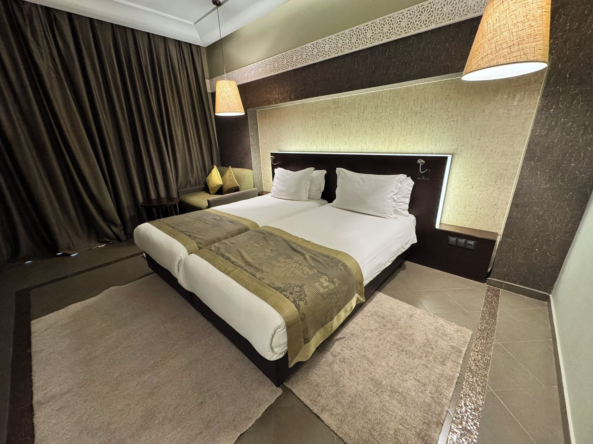 A hotel room with a large bed, two bedside tables, a headboard, a curtain, and two wall-mounted lamps.