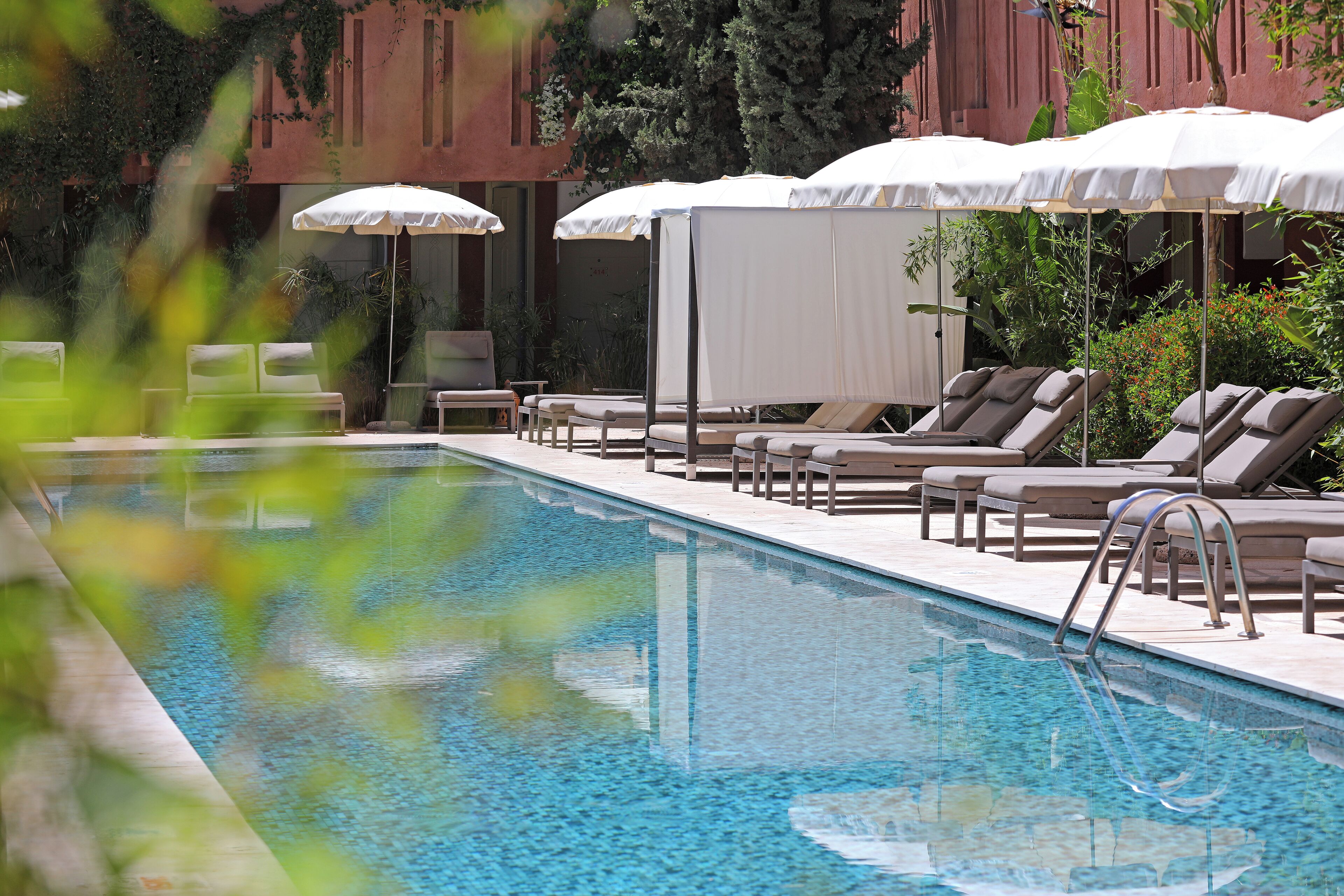 A swimming pool with lounge chairs and umbrellas, surrounded by greenery and a building with a red facade.
