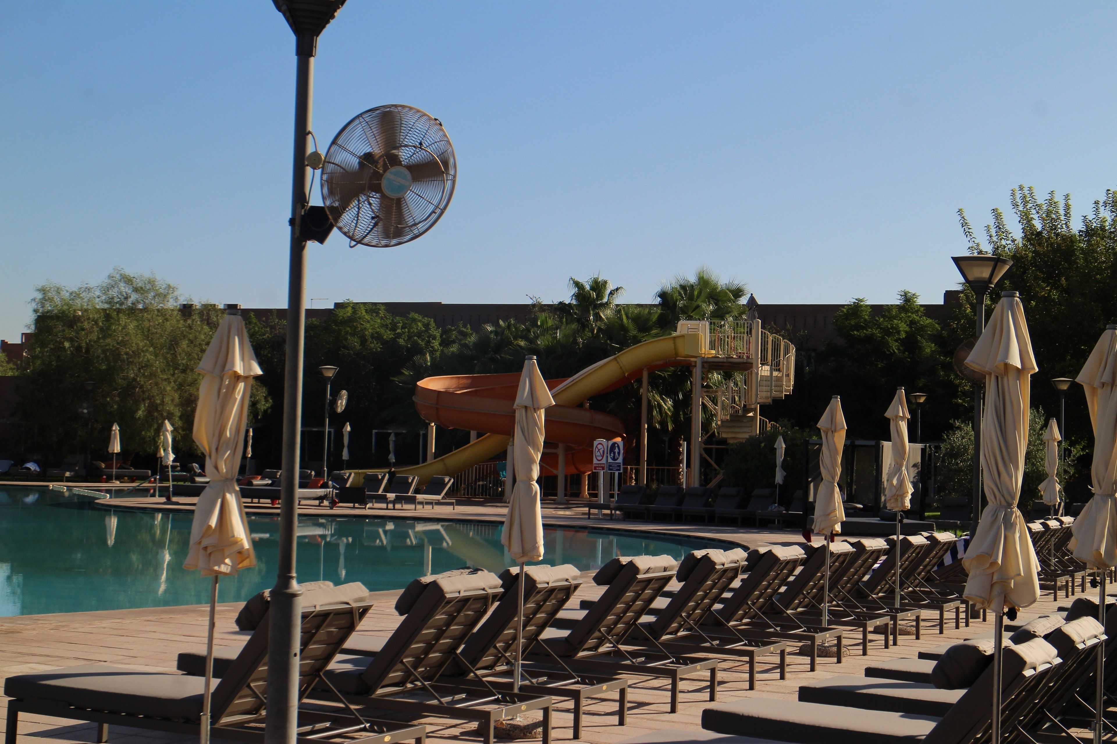 Zona de piscina con sillones reclinables, sombrillas, un ventilador y un tobogán de agua.