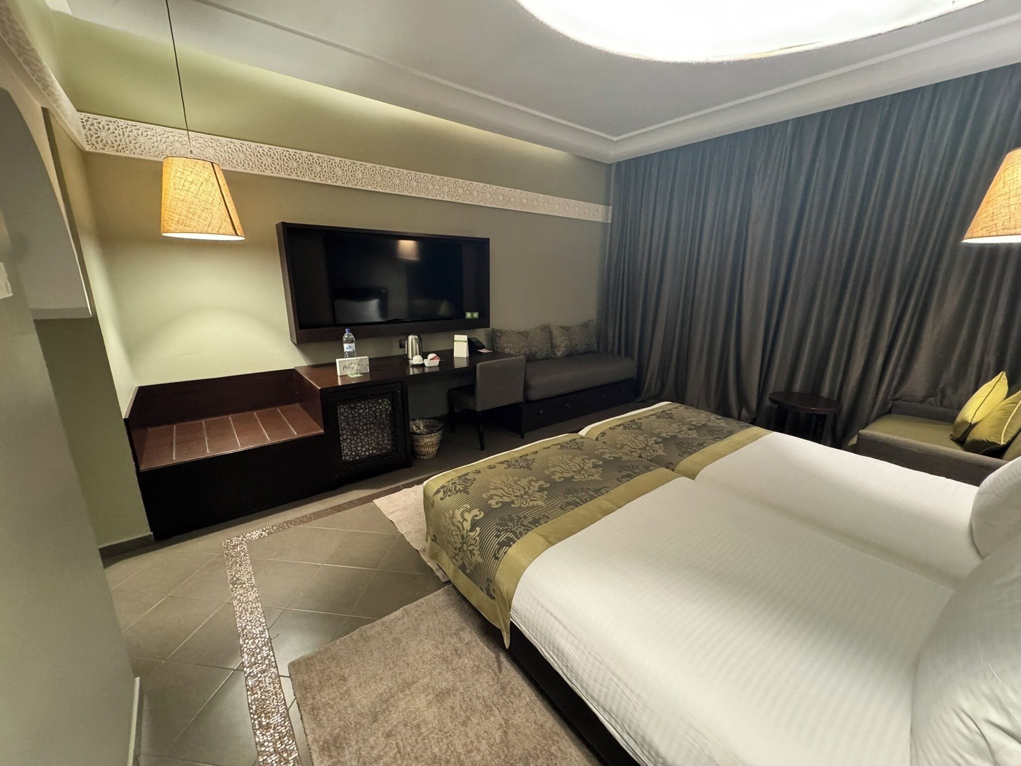 A hotel room with a large bed, a desk, a flat-screen TV, and a seating area with curtains.