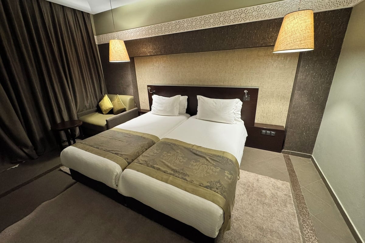 Una habitación de hotel con una cama grande, dos mesitas de noche, una silla y una lámpara.