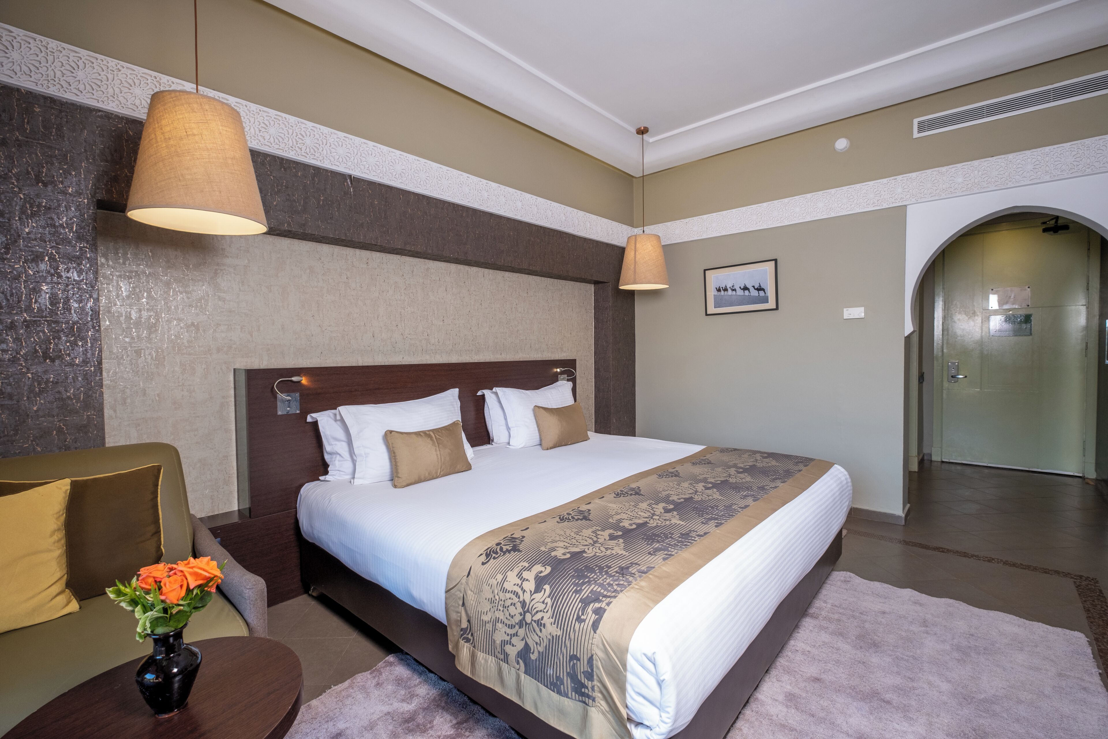 A hotel room with a large bed, a wooden headboard, a small table with a vase of flowers, and a door leading to a bathroom.